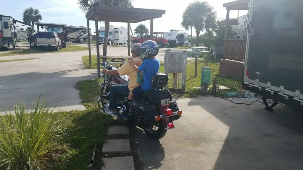 Photo of a man and child on a motorcycle inside the Florida RV Beach resort Bryn Mawr. There are several RVs and palm trees around